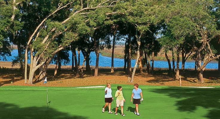 Three women walking on a lush greenway with large oak trees and a waterway behind them at The Landings Golf & Athletic Club.