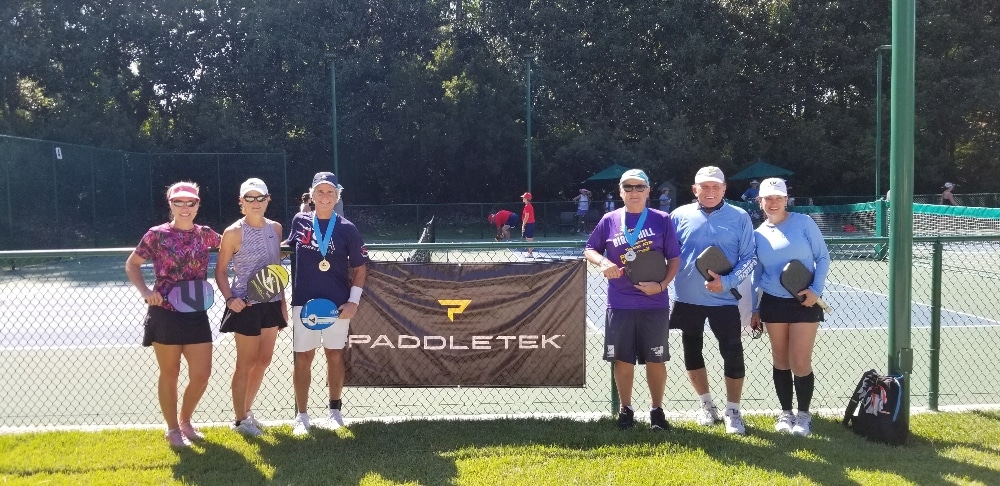 Six pickleball players gather around a banner at the Pickle-Palooza tournament.