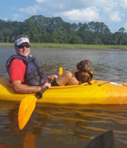 A man wearing a visor and lifejacket is joined by his dachshund dog kayaking in a yellow kayak on the water.