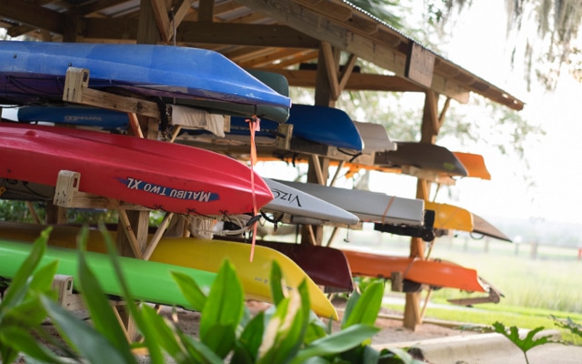 Several blue, green or red kayaks are stacked under an awning awaiting rental.