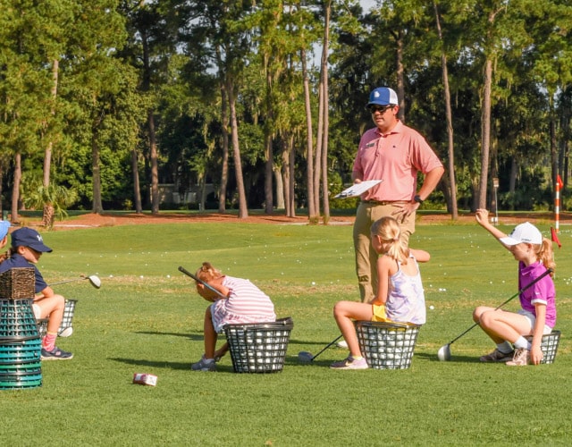 Golf instructor talking to three young girls on a golf course who are sitting on buckets of golf balls.