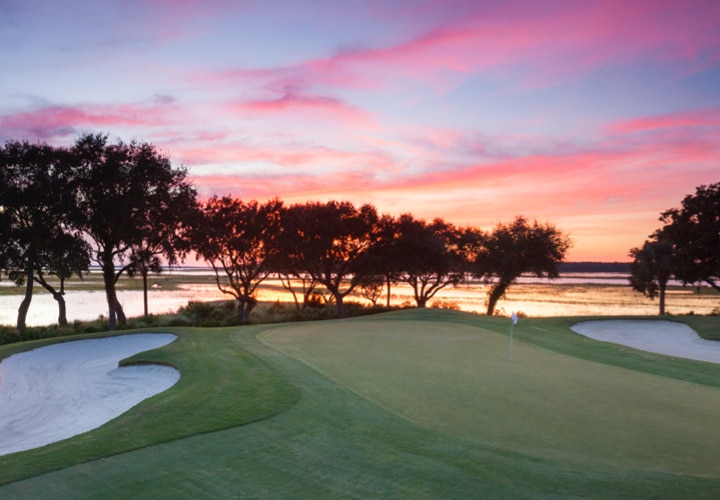 A row of trees on the green are backlit by a vibrant pink sunset on the Terrapin Point golf course, designed by Willard C. Byrd.