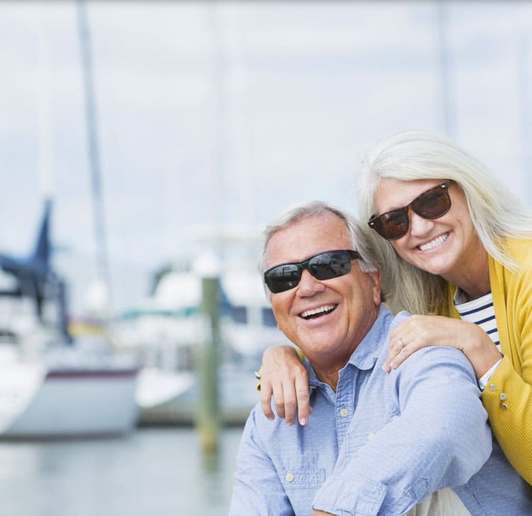 Retired man and woman, both wearing sunglasses, smile for a photo with the boats taken at the marina.