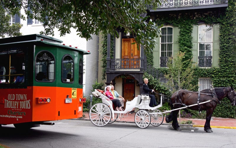 A green and red Old Town Trolley Tours trolley passes by a horse-drawn carriage ferrying tourists through the  downtown Savannah streets.