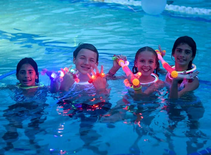 Four children bobbing in the water show off their glow-in-the dark jewelry at a Friday Swim Night at Franklin Creek Pool.