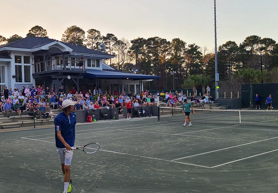 Two men finish up a tennis match as a group of fans gather outside the clubhouse.
