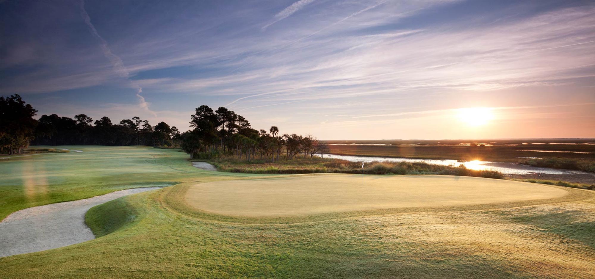 A sand trap lies to the left and a marsh to the right of his green at sunset on the Palmetto golf course.