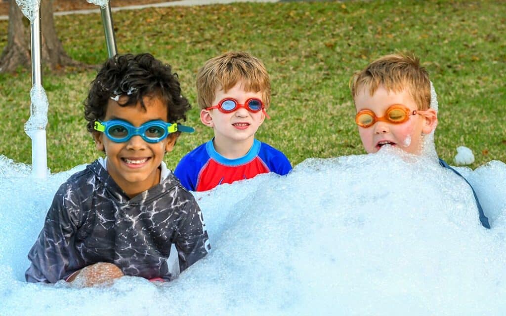 Three boys wearing goggles sit and smile from a pool of foam during a kids night out event.