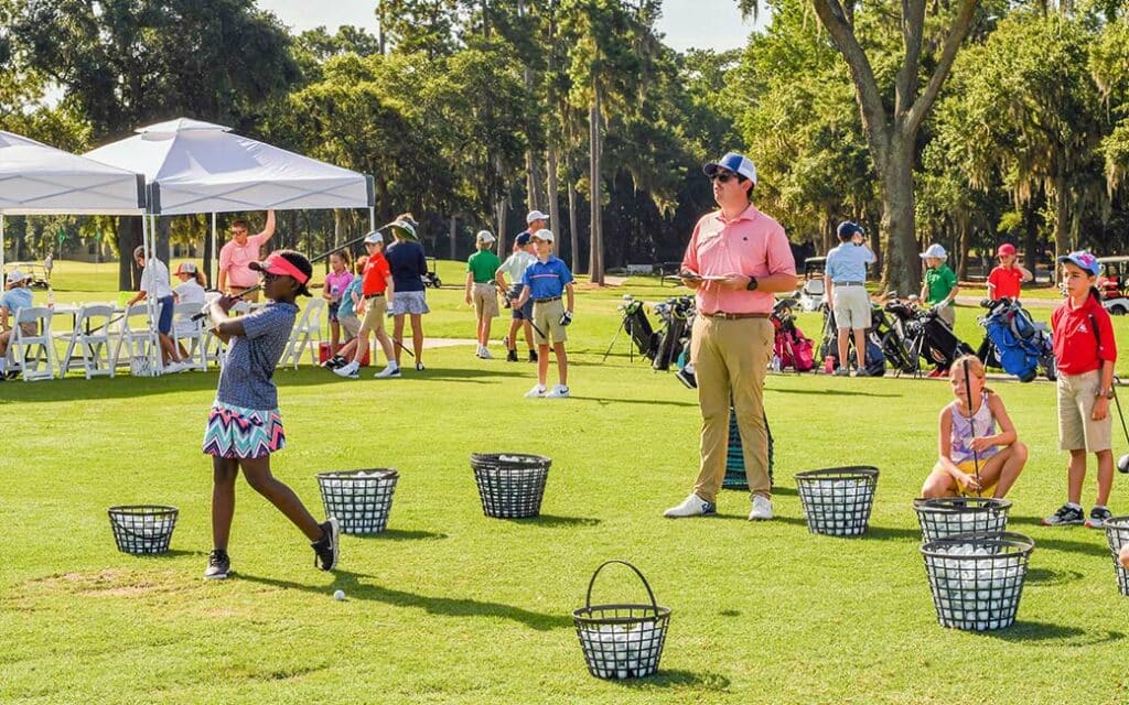 An instructor teaches a group of kids how to golf,  each complete with a bucket of balls.